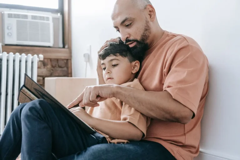 A father sitting on the floor at home, reading with his young son on his lap