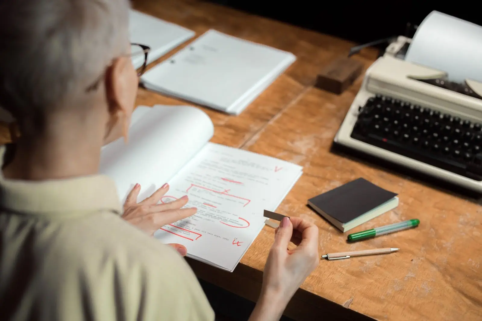 Woman correcting a printed draft with a red pen beside a typewriter