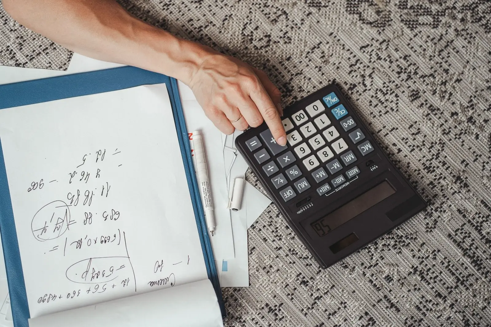 Hand on a calculator beside handwritten sums